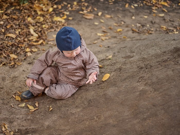 Ein Kleinkind in brauner, gepunkteter Outdoor-Kleidung und blauer Mütze sitzt auf einem erdigen Boden im Freien und betrachtet ein Herbstblatt in seiner Hand; rundherum liegen weitere gelbe und braune Blätter.