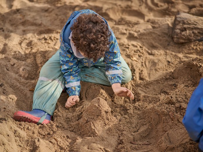 Ein Kind mit lockigem Haar sitzt im Sand und spielt mit den Händen; es trägt eine gemusterte blaue Jacke und grüne Matschhose, vor ihm ist ein kleiner Sandhügel geformt, der Boden besteht aus lockerem Sand.