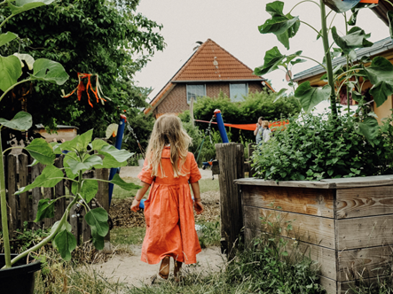 Ein Mädchen in einem orangefarbenen Kleid läuft von hinten durch einen üppig bepflanzten Garten. Rechts steht ein Holzhochbeet mit Pflanzen, links wachsen große Sonnenblumen. Im Hintergrund ist ein Haus mit rotem Ziegeldach sowie weitere Kinder und ein Spielgerüst zu sehen.
