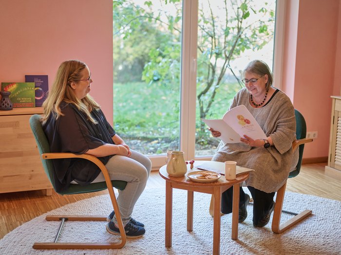Zwei Waldorferzieherinnen sitzen in einem freundlichen Gespräch zusammen, eine zeigt der anderen eine Broschüre – eine kollegiale Besprechung in einem warm gestalteten Raum der Einrichtung.