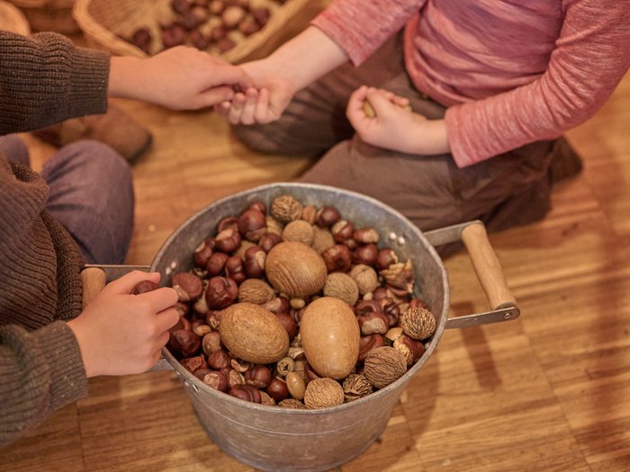 Zwei Kinder sitzen auf einem Holzboden und halten gemeinsam einen Metalleimer voller verschiedener Nüsse, darunter Kastanien, Haselnüsse und Walnüsse. Im Hintergrund sind weitere Körbe mit Nüssen zu sehen.