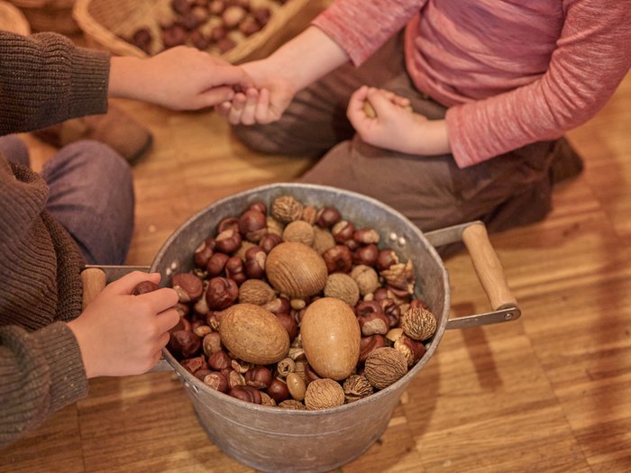 Zwei Kinder sitzen auf einem Holzboden und halten gemeinsam einen Metalleimer voller verschiedener Nüsse, darunter Kastanien, Haselnüsse und Walnüsse. Im Hintergrund sind weitere Körbe mit Nüssen zu sehen.