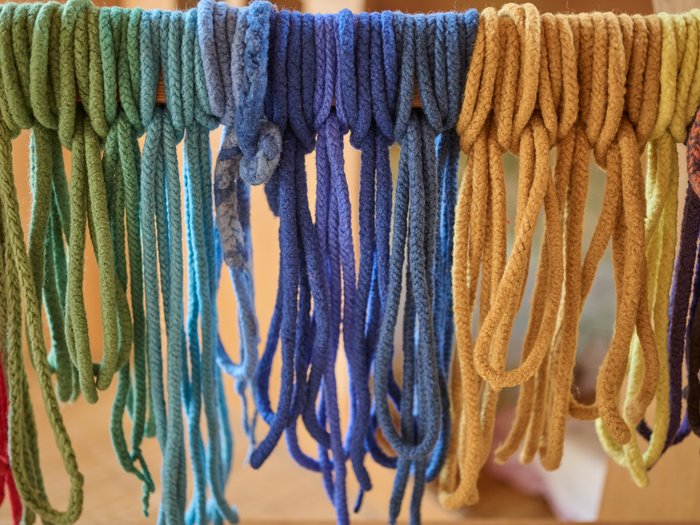 Mehrere dicke, weiche Woll- oder Garnstränge hängen in Schlaufen über einer Stange; sie sind nach Farben sortiert und verlaufen von Grün- und Türkistönen über Blau bis zu Gelb- und Ockertönen vor einem unscharfen Hintergrund.