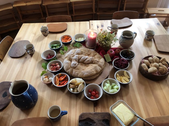 Festlich gedeckter Holztisch mit einem großen runden Michaelibrot in der Mitte, umgeben von kleinen Schälchen mit frischem Obst, Gemüse und saisonalen Zutaten sowie einer brennenden Kerze und einem Blumenstrauß.
