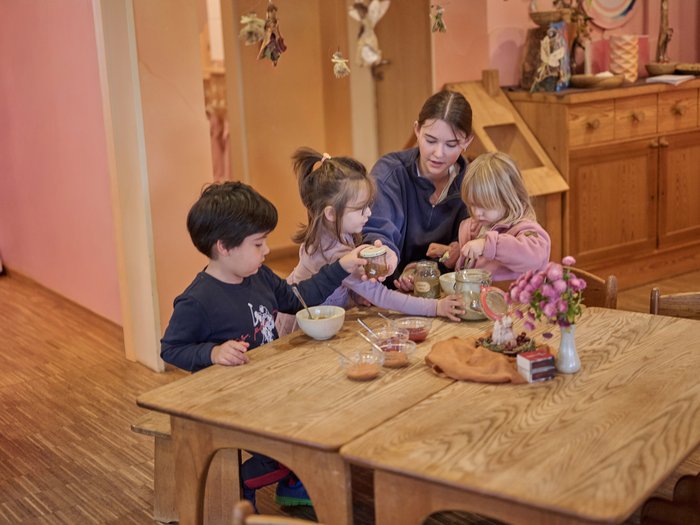 Eine junge Waldorferzieherin sitzt mit drei Kleinkindern an einem massiven Holztisch und untersucht gemeinsam mit ihnen verschiedene Einmachgläser mit Naturmaterialien.