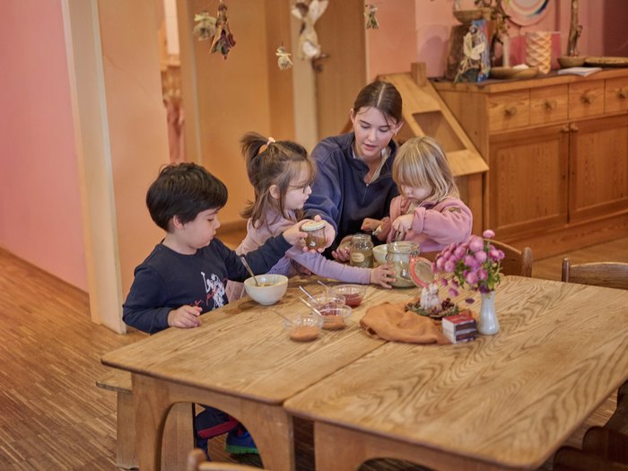 Eine junge Waldorferzieherin sitzt mit drei Kleinkindern an einem massiven Holztisch und untersucht gemeinsam mit ihnen verschiedene Einmachgläser mit Naturmaterialien.