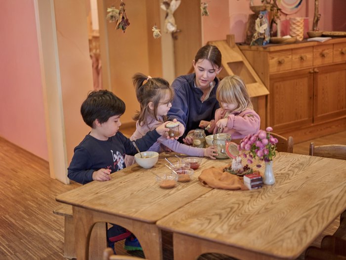 Eine junge Waldorferzieherin sitzt mit drei Kleinkindern an einem massiven Holztisch und untersucht gemeinsam mit ihnen verschiedene Einmachgläser mit Naturmaterialien.