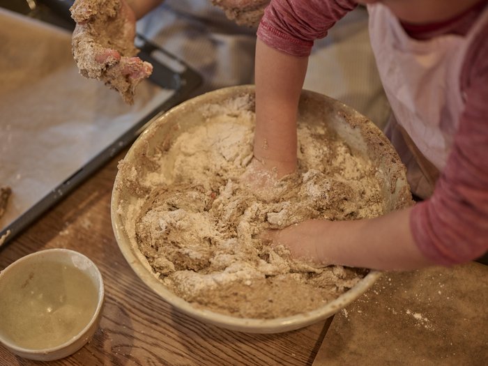 Kinderhände kneten eine mehlige Teigmasse in einer großen Schüssel; daneben stehen eine kleine Keramikschale und ein Backblech, alles auf einem Holztisch in einem Innenraum.