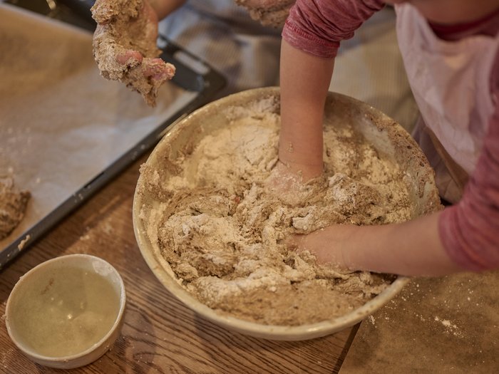 Kinderhände kneten eine mehlige Teigmasse in einer großen Schüssel; daneben stehen eine kleine Keramikschale und ein Backblech, alles auf einem Holztisch in einem Innenraum.