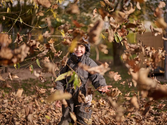 Ein lachendes Kind in dunkler Outdoor-Kleidung steht im Freien und wirft Herbstlaub in die Luft; um das Kind herum fliegen braune und gelbe Blätter, im Hintergrund sind Bäume und ein herbstlicher Garten zu sehen.