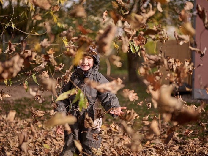 Ein lachendes Kind in dunkler Outdoor-Kleidung steht im Freien und wirft Herbstlaub in die Luft; um das Kind herum fliegen braune und gelbe Blätter, im Hintergrund sind Bäume und ein herbstlicher Garten zu sehen.